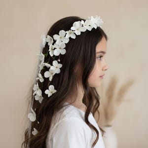 May include: A white floral hair crown and cascading floral accents adorn dark brown hair. The flowers are white and delicate, creating a natural, elegant look. The person is wearing a white top.