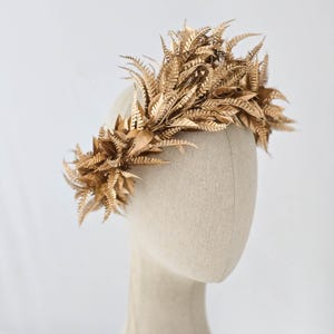 May include: A golden headpiece featuring a crown of stylized leaves and fern fronds. The headpiece is displayed on a mannequin head. The leaves and fronds are painted in a metallic gold color.