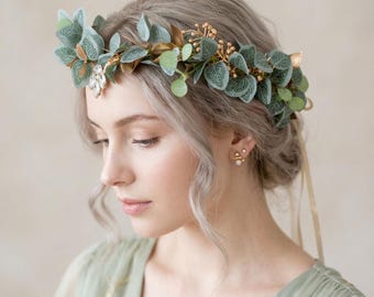 May include: A floral crown featuring sage green eucalyptus leaves, small gold-toned berries, and a central clear crystal embellishment. The headpiece is secured with long, thin ribbons trailing from the back, creating a delicate botanical accessory.