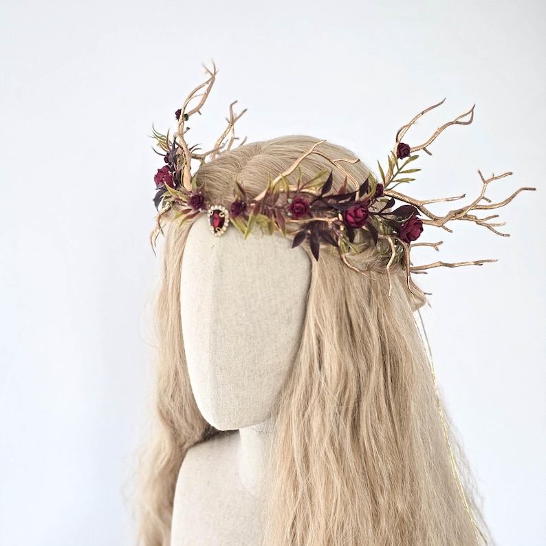 May include: A handcrafted headpiece featuring a gold-toned, twig-like frame adorned with burgundy roses, dark purple leaves, and a central red jewel. The crown is designed for a fantasy or woodland theme, perfect for special events.