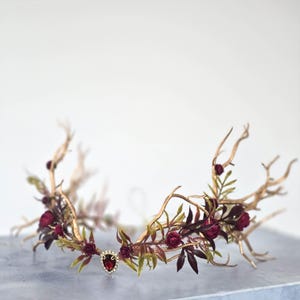May include: A gold-toned crown with a central red jewel and surrounding burgundy roses and leaves. The crown's design incorporates branches and foliage, creating a natural, elegant aesthetic. The headpiece is suitable for special occasions.