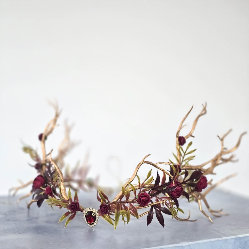 May include: A gold-toned crown with a central red jewel and surrounding burgundy roses and leaves. The crown's design incorporates branches and foliage, creating a natural, elegant aesthetic. The headpiece is suitable for special occasions.