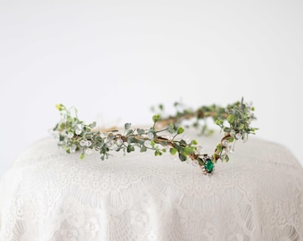 May include: A delicate floral crown featuring faux greenery, clear crystal beads, and a central emerald-colored jewel. The crown is displayed on a white lace surface, perfect for weddings or special occasions.