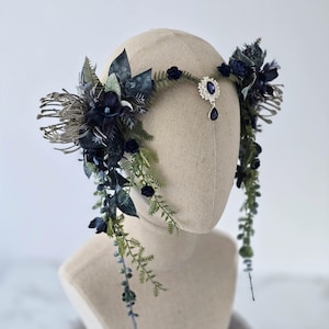 Pode incluir: Uma tiara floral azul escuro com uma joia central e uma l&aacute;grima pendurada. A tiara apresenta flores azuis escuras, folhas verdes e detalhes cinzentos. A tiara &eacute; exibida em uma cabe&ccedil;a de manequim.