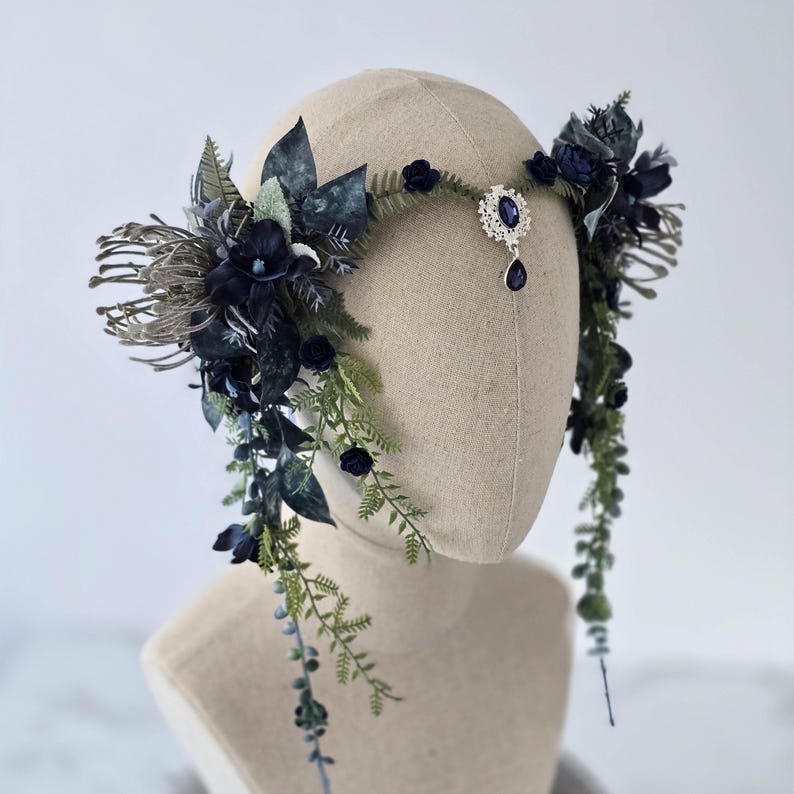 Pode incluir: Uma tiara floral azul escuro com uma joia central e uma l&aacute;grima pendurada. A tiara apresenta flores azuis escuras, folhas verdes e detalhes cinzentos. A tiara &eacute; exibida em uma cabe&ccedil;a de manequim.