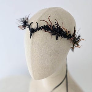 May include: A gothic-style crown with a black bird skull and crescent moon design. The crown is decorated with black and brown dried foliage, creating a dramatic headpiece. The crown is displayed on a mannequin head.