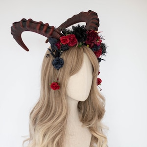 May include: A headpiece featuring large, curved, dark red horns. The horns are set on a crown of black and red flowers and dark blue accents. The headpiece is displayed on a mannequin head with long, wavy blonde hair.
