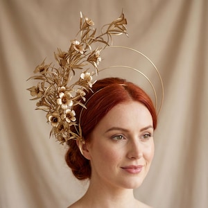 May include: A gold-coloured headpiece with floral and halo elements. The headpiece features a cluster of flowers and leaves on one side, with concentric circles above the head. Worn by a person with red hair, the headpiece is an elegant accessory.