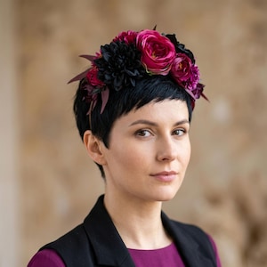 Könnte beinhalten: Eine Frau mit einem Blumenkranz aus dunkelrosa Rosen, schwarzen Dahlien und burgunderfarbenen Akzenten. Der Kranz sitzt auf kurzem, dunklem Haar. Sie trägt ein lila Oberteil und einen schwarzen Blazer.