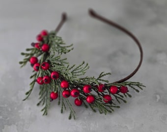May include: A thin brown headband adorned with a cluster of artificial green cedar foliage and small, round, bright red berries. The decorative arrangement is attached to one side of the band, featuring textured evergreen needles and glossy red fruit.
