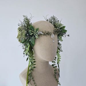 Puede incluir: Una corona floral artesanal con una variedad de follaje verde y gris, incluyendo hojas y ramitas. Un adorno central plateado con un cristal colgante añade un toque de elegancia. La corona se exhibe en una cabeza de maniquí.