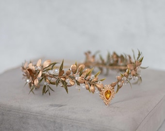 May include: A gold leaf and crystal bridal crown with a single amber gemstone accent. The crown is made of delicate gold leaves and branches, with clear crystals scattered throughout. The crown is perfect for a bohemian or woodland wedding.