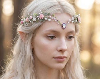 May include: A delicate floral crown featuring pink and white fabric flowers, green leaves, and faux pearls. The crown has a central oval-shaped gemstone and is designed for a fairytale or fantasy aesthetic. The crown is worn on the head.