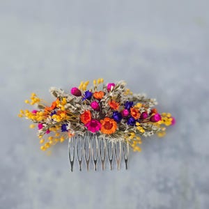 Puede incluir: Un vibrante peine floral con una variedad de flores secas coloridas. El peine incluye flores amarillas, naranjas, rosas y moradas, dispuestas sobre una base de peine plateada. Las flores están agrupadas, creando una apariencia completa y texturizada.
