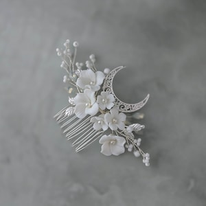 Op de afbeelding: Een zilveren haar kam met een maan ontwerp en delicate witte bloemen met parel accenten. De kam heeft een reeks fijne tanden en is versierd met kleine, parel bezette takjes en bladeren. Een romantisch haaraccessoire.