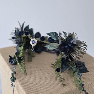 Pode incluir: Uma coroa floral azul escuro com um pendente central prateado com uma pedra preciosa azul. A coroa &eacute; adornada com rosas azul escuro, folhas verdes e vegeta&ccedil;&atilde;o rasteira, criando um design elegante inspirado na natureza.