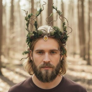 Peut inclure: Une couronne inspirée de la nature, ornée de branches de conifères, de pommes de pin et de brindilles. Un bijou doré est centré sur le front. La couronne est portée par une personne avec une barbe et de longs cheveux, sur fond de forêt flou.