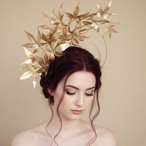 May include: A gold-coloured headpiece with maple leaf designs. The leaves are arranged around a circular frame, creating a crown-like effect. The headpiece is worn by a person with dark red hair, against a neutral background.