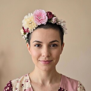Könnte beinhalten: Ein Blumenkranz mit rosa, cremefarbenen und burgunderfarbenen Blumen. Der Kranz wird auf dem Kopf einer Person getragen. Die Person trägt ein Oberteil mit Blumenmuster. Der Hintergrund ist einfarbig.