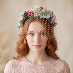 Peut inclure: Un accessoire de tête floral avec un voile de type voilette blanc, orné d'un arrangement de fleurs artificielles dans des tons rose, bleu et crème, agrémenté de feuilles vertes et de baies sombres, sur un vêtement en dentelle rose pâle.