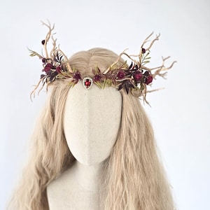 May include: A decorative headpiece featuring a crown design with gold-colored branches, burgundy flowers, and green leaves. A red jewel is centered on the forehead. The headpiece is displayed on a mannequin head with long, wavy blonde hair.