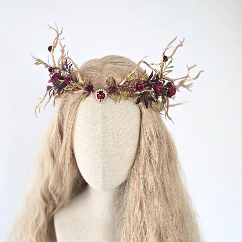 May include: A decorative headpiece featuring a crown design with gold-colored branches, burgundy flowers, and green leaves. A red jewel is centered on the forehead. The headpiece is displayed on a mannequin head with long, wavy blonde hair.