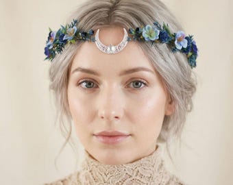 May include: A floral headpiece featuring a silver-toned crescent moon charm with a central teardrop-shaped stone. The band is adorned with an arrangement of small blue and teal fabric flowers, dark green leaves, and sprigs of foliage.