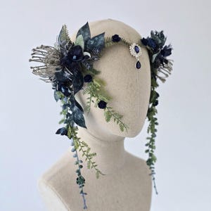 Pode incluir: Uma tiara floral azul escuro com vegeta&ccedil;&atilde;o em cascata e uma joia central. A tiara apresenta flores azul escuro, folhas verdes e um adorno prateado com uma joia em forma de gota. Apresentada em uma cabe&ccedil;a de manequim.