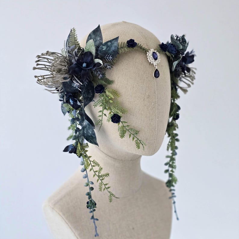 Pode incluir: Uma tiara floral azul escuro com vegeta&ccedil;&atilde;o em cascata e uma joia central. A tiara apresenta flores azul escuro, folhas verdes e um adorno prateado com uma joia em forma de gota. Apresentada em uma cabe&ccedil;a de manequim.