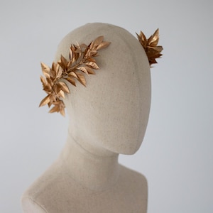May include: A gold-colored leaf crown, likely for a special occasion, is displayed on a mannequin head. The leaves are arranged in a symmetrical pattern, with a few leaves on the side and top of the head. The background is a plain white.