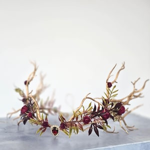 May include: A gold-toned crown with a branch-like design, adorned with burgundy roses and green leaves. A central jewel adds a touch of elegance. This headpiece is ideal for weddings or special occasions.
