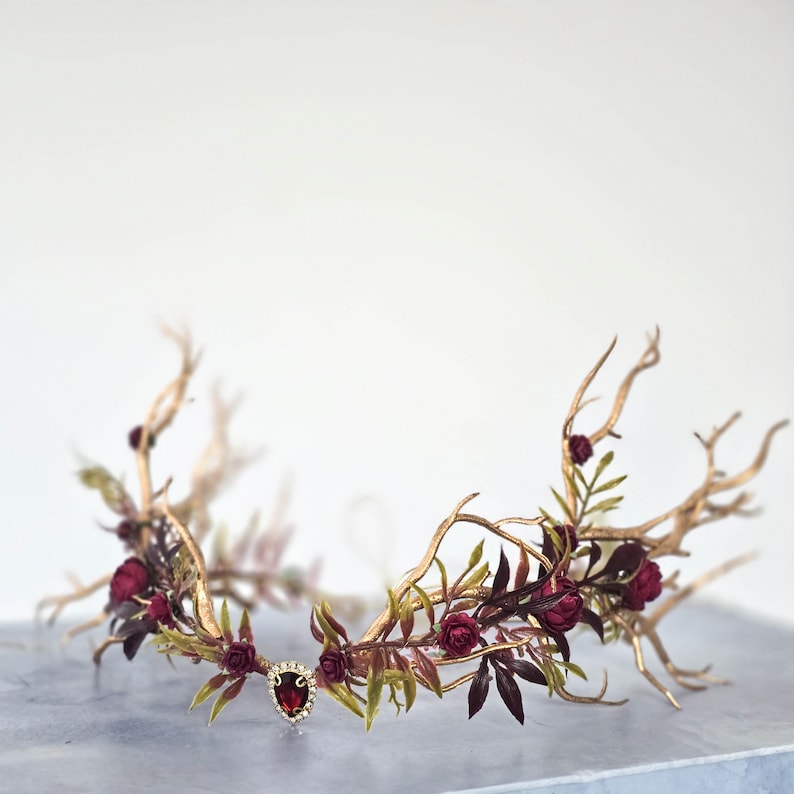 May include: A gold-toned crown with a branch-like design, adorned with burgundy roses and green leaves. A central jewel adds a touch of elegance. This headpiece is ideal for weddings or special occasions.