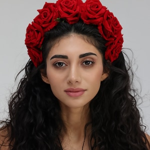May include: A woman with long, dark curly hair wearing a red rose flower crown.