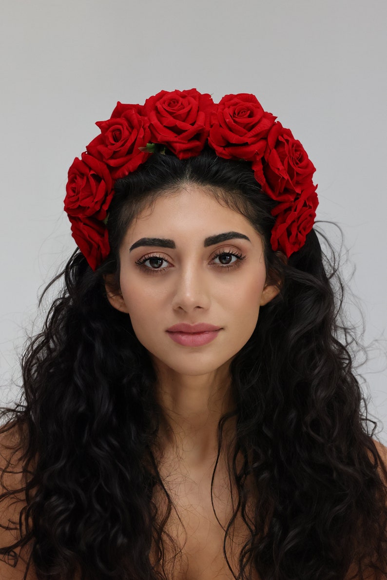 May include: A woman with long, dark curly hair wearing a red rose flower crown.