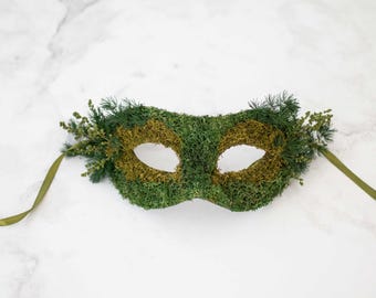 Puede incluir: Una máscara de masquerade verde y dorada hecha de musgo y vegetación. La máscara tiene una cinta verde.