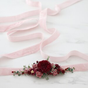 May include: A pink velvet ribbon with a burgundy and blush floral embellishment. The flowers are made of fabric and are arranged in a cluster. The ribbon is tied in a bow.