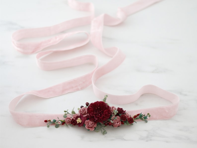 May include: A pink velvet ribbon with a burgundy and blush floral embellishment. The flowers are made of fabric and are arranged in a cluster. The ribbon is tied in a bow.