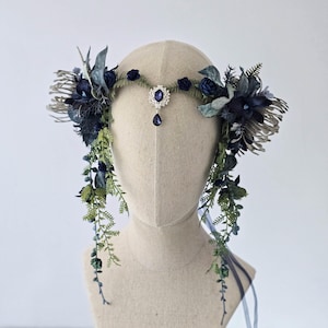 Pode incluir: Uma coroa floral azul escuro com uma joia central e detalhes pendurados. A pe&ccedil;a de cabe&ccedil;a apresenta v&aacute;rias tonalidades de flores azuis, folhagem verde e detalhes prateados. Fitas penduradas nas laterais. Uma pe&ccedil;a de cabe&ccedil;a decorativa para ocasi&otilde;es especiais.