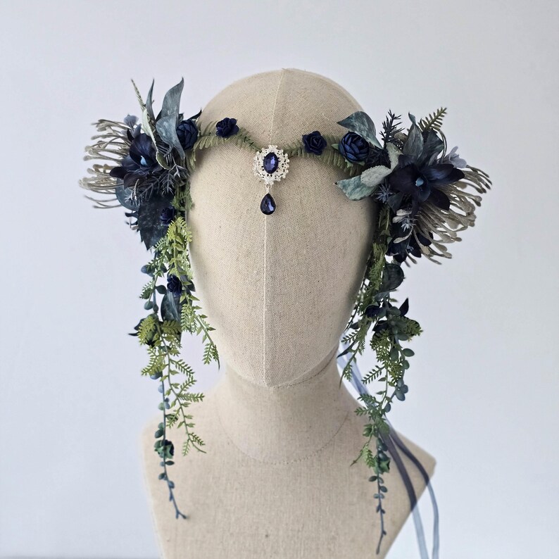 Pode incluir: Uma coroa floral azul escuro com uma joia central e detalhes pendurados. A pe&ccedil;a de cabe&ccedil;a apresenta v&aacute;rias tonalidades de flores azuis, folhagem verde e detalhes prateados. Fitas penduradas nas laterais. Uma pe&ccedil;a de cabe&ccedil;a decorativa para ocasi&otilde;es especiais.