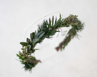 May include: A handcrafted floral crown featuring a white wire base adorned with an assortment of preserved greenery. The crown includes dark green leaves, sprigs of dried flowers, and various textures, perfect for weddings or special occasions.