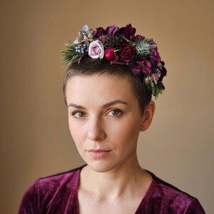 Könnte beinhalten: Ein Blumenkranz mit einer Vielzahl von Blumen in Lila-, Burgunder- und Rosatönen, akzentuiert mit grünem Laub und kleinen blauen Beeren. Der Kranz wird auf dem Kopf getragen.