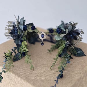 Pode incluir: Uma coroa floral artesanal com folhas e flores azuis escuras e verdes. A coroa tem um pendente central de joia com uma gema pendurada em forma de gota. A coroa &eacute; projetada com uma mistura de texturas e tons de azul e verde.