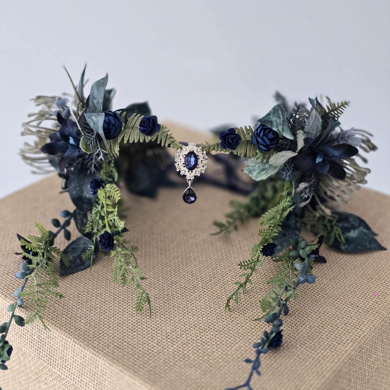 Pode incluir: Uma coroa floral artesanal com folhas e flores azuis escuras e verdes. A coroa tem um pendente central de joia com uma gema pendurada em forma de gota. A coroa &eacute; projetada com uma mistura de texturas e tons de azul e verde.