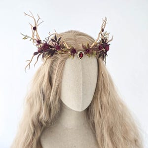 May include: A handcrafted headpiece featuring a crown design with faux antlers, burgundy roses, and green leaves. A central red jewel adds a focal point. The headpiece is displayed on a mannequin head with long, wavy blonde hair.