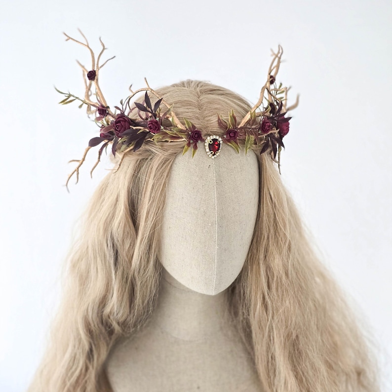 May include: A handcrafted headpiece featuring a crown design with faux antlers, burgundy roses, and green leaves. A central red jewel adds a focal point. The headpiece is displayed on a mannequin head with long, wavy blonde hair.