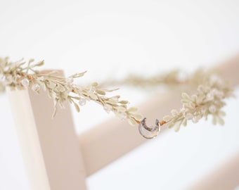 May include: A delicate floral crown featuring light green leaves and clear crystal beads. A small, iridescent crescent moon charm is centered on the crown. The crown is designed for special occasions.