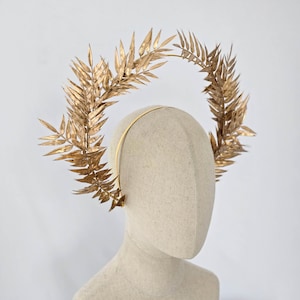 May include: A gold-colored headpiece featuring a circular design of stylized leaves. The leaves are arranged in a wreath-like pattern, attached to a thin, gold headband. The headpiece is displayed on a mannequin head against a white background.