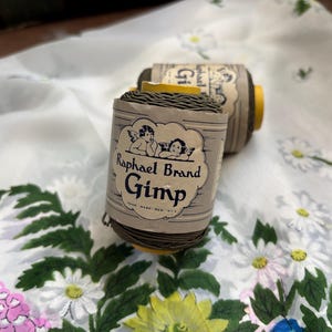 May include: Two spools of Raphael Brand Gimp thread, with a vintage label featuring cherubs. The spools are wrapped in a light gray paper with a yellow base. The thread is a dark olive green color.