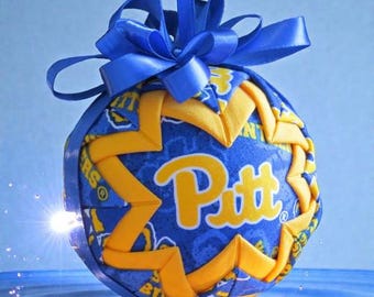 May include: A handcrafted ornament featuring a blue and gold design with the word "Pitt". The ornament is round, with a blue ribbon for hanging and a decorative bow at the top. The design incorporates a star pattern.