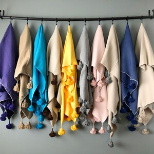 Puede incluir: Diez mantas de colores con ribete de pompones cuelgan de una barra de metal negro. Las mantas son en tonos de morado, beige, azul, amarillo, gris, rosa y blanco.